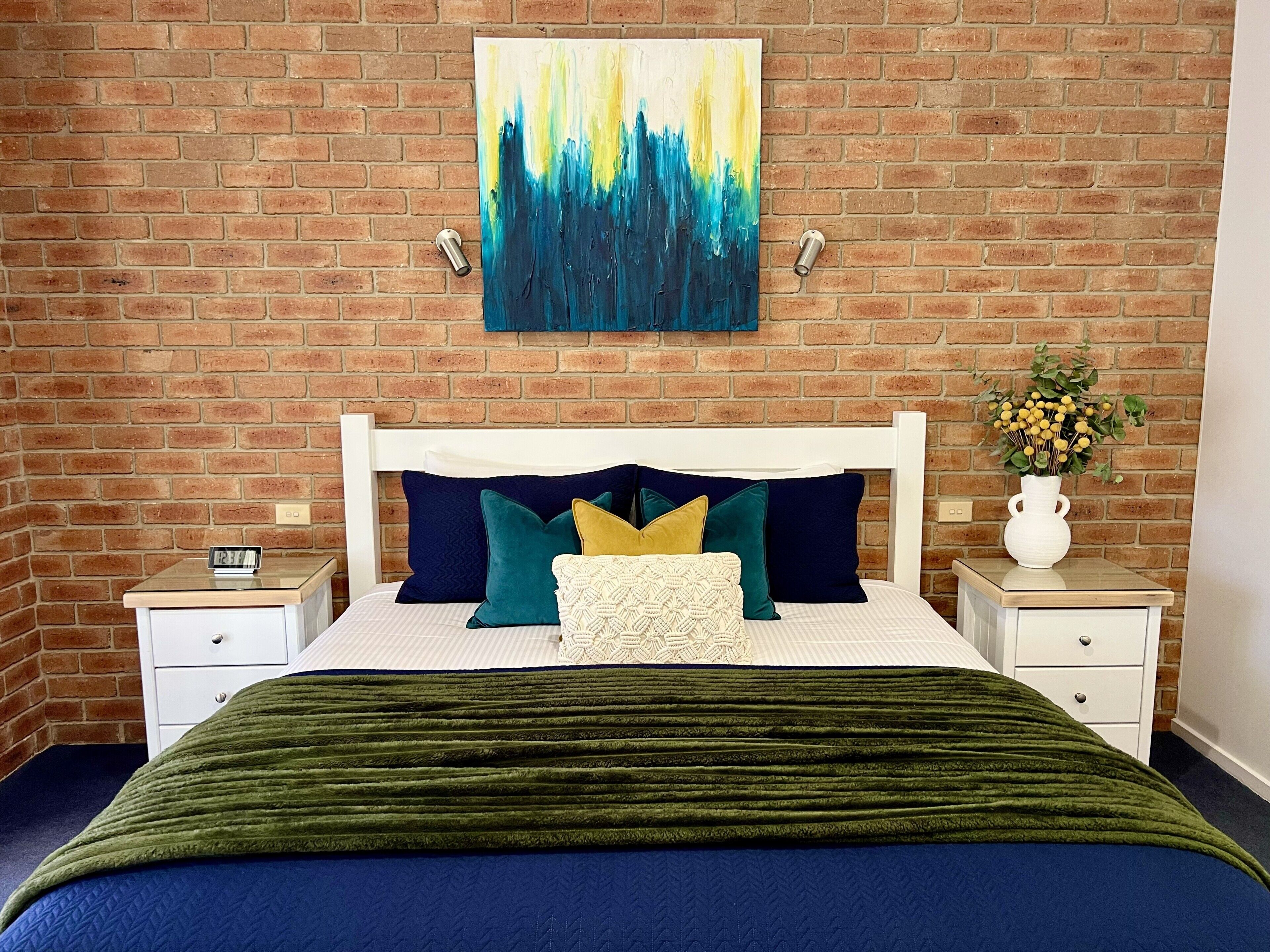A bedroom with a brick wall, a bed with a white headboard, multiple pillows, a green blanket, a nightstand with a clock, and a vase with flowers.