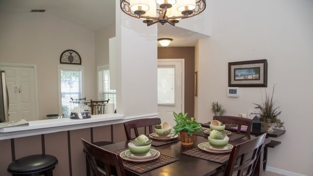 A dining area with a wooden table set for four, surrounded by chairs. A kitchen counter with a refrigerator and a clock is visible in the background. - Ly53820 - Indian Ridge - 3 Bed 2 Baths Villa (Kissimmee)