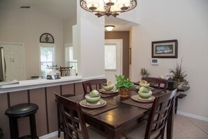 A dining area with a wooden table set for four, surrounded by chairs. A kitchen counter with a refrigerator and a clock is visible in the background. - Ly53820 - Indian Ridge - 3 Bed 2 Baths Villa (Kissimmee)