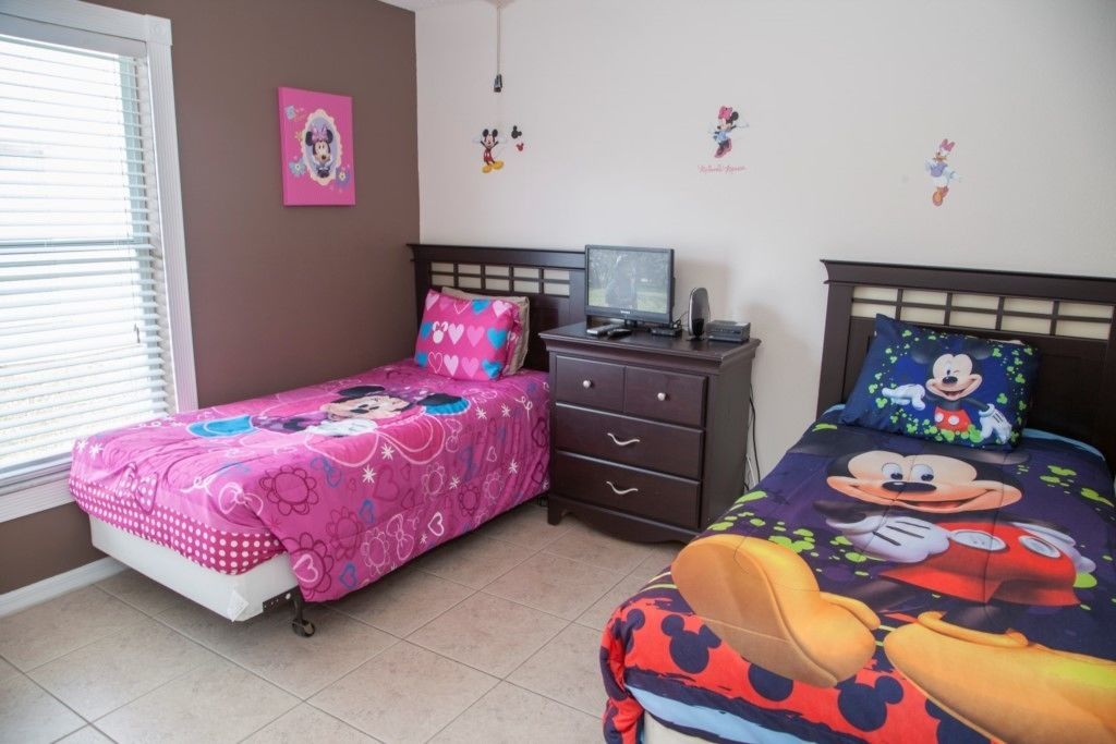 A bedroom with two beds, one with a pink Minnie Mouse theme and the other with a Mickey Mouse theme. There is a dresser with a computer and a small table.