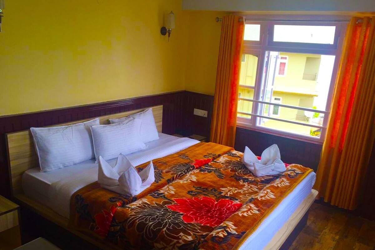 A hotel room with a bed, a nightstand, a window with curtains, and a view of buildings outside.