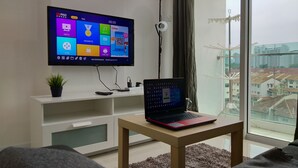 A modern living room with a large TV mounted on the wall, a laptop on a small table, and a view of the city through a large window. - Suria Next To Desa ParkCity (Kuala Lumpur)