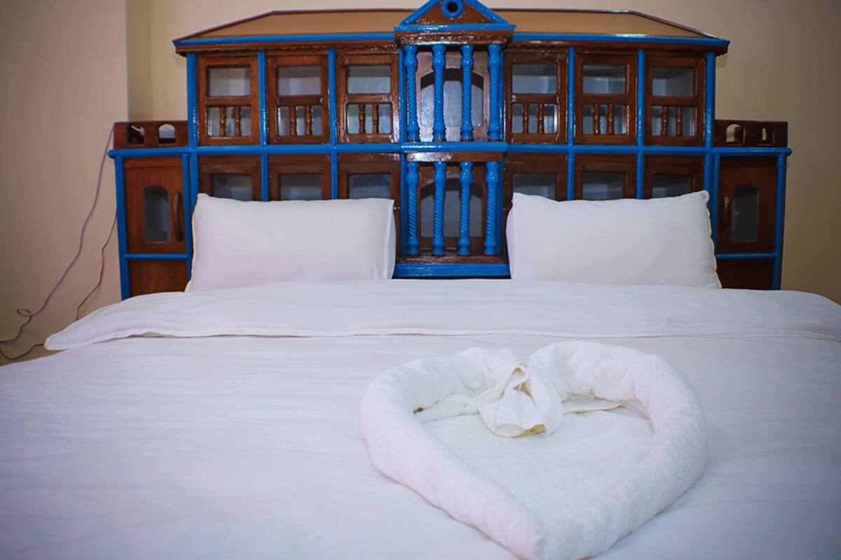 A bed with a white comforter, two white pillows, and a heart-shaped towel on the bed.
