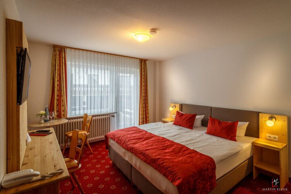 Une chambre d’hôtel avec un lit, un bureau, une chaise, un radiateur et une fenêtre avec des rideaux.