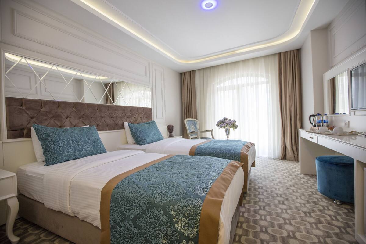 Quarto de hotel com duas camas, uma escrivaninha, uma cadeira, toucador e uma janela grande com cortinas.