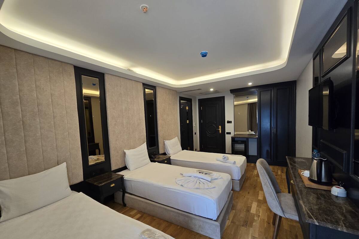 Quarto de hotel com três camas, uma escrivaninha, uma cadeira, uma televisão e um espelho.
