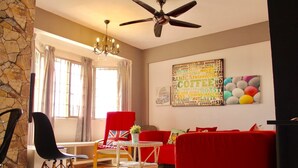 A living room with a ceiling fan, a chandelier, a red sofa, a red chair, a small table with a plant, and a word cloud artwork on the wall. - Aman Dua Apartment by SYNC (Batu Caves)