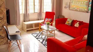 A living room with a red sofa, a red armchair, a small table with a potted plant, and a bench. There is a word cloud on the wall and a stone wall on one side. - Aman Dua Apartment by SYNC (Batu Caves)
