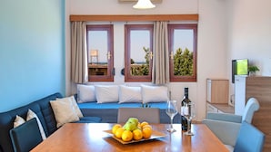 A modern room with a dining table set with fruit and wine, a sofa with cushions, a TV, and a view of the outdoors through large windows. - Island Suites (Apokoronas)