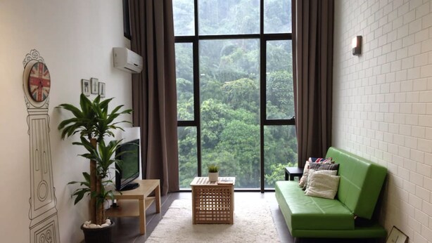 Una sala de estar con un ventanal grande, un sofá verde, una mesa de madera, una planta en maceta y un reloj fijado a la pared.