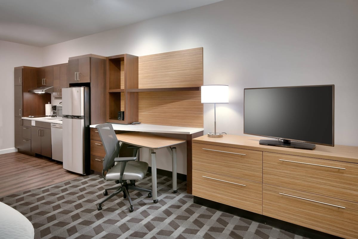 A modern hotel room with a kitchenette, a desk, a chair, a flat-screen TV, and a lamp.