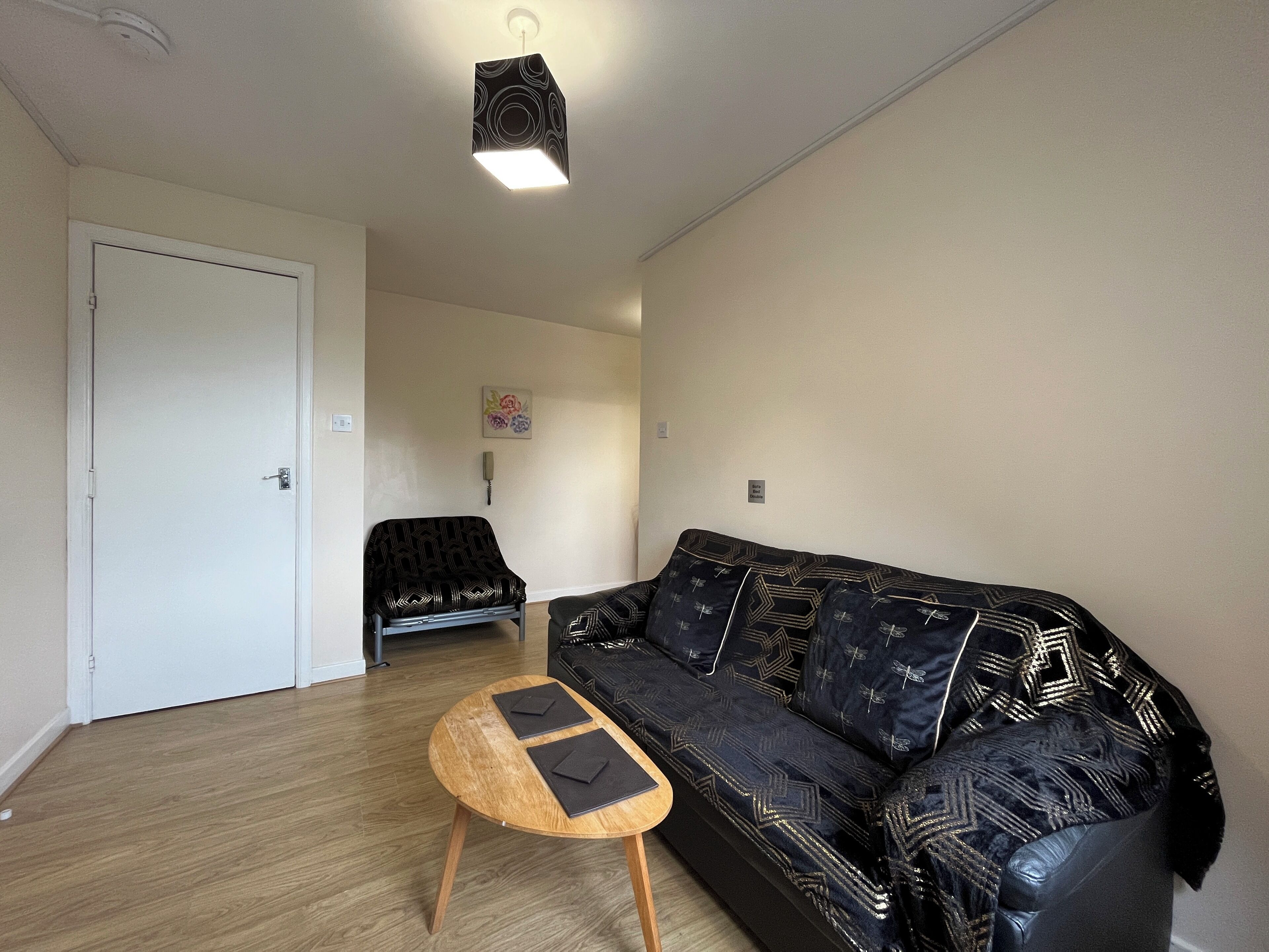A living room with a black patterned sofa, a wooden coffee table with two black placemats, a white door, and a ceiling light fixture.