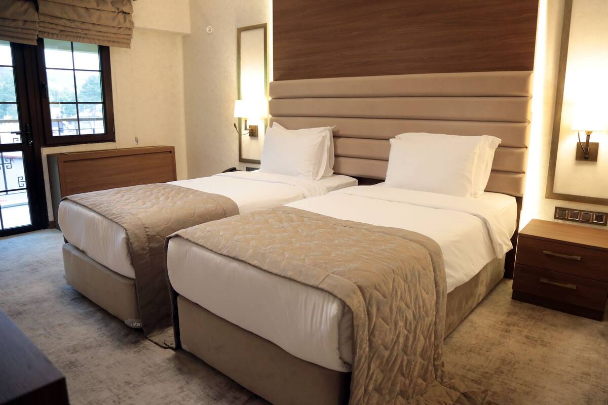 A hotel room with two beds, a wooden headboard, a nightstand, and a window with a view.