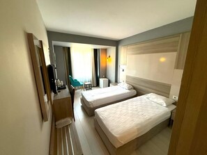 Habitación de hotel con dos camas individuales, un escritorio con silla, un televisor y un ventanal con cortinas.