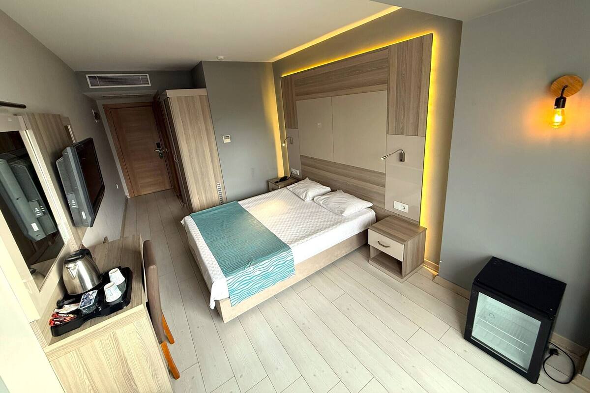 A modern hotel room with a bed, bedside table, TV, and a kitchenette area.