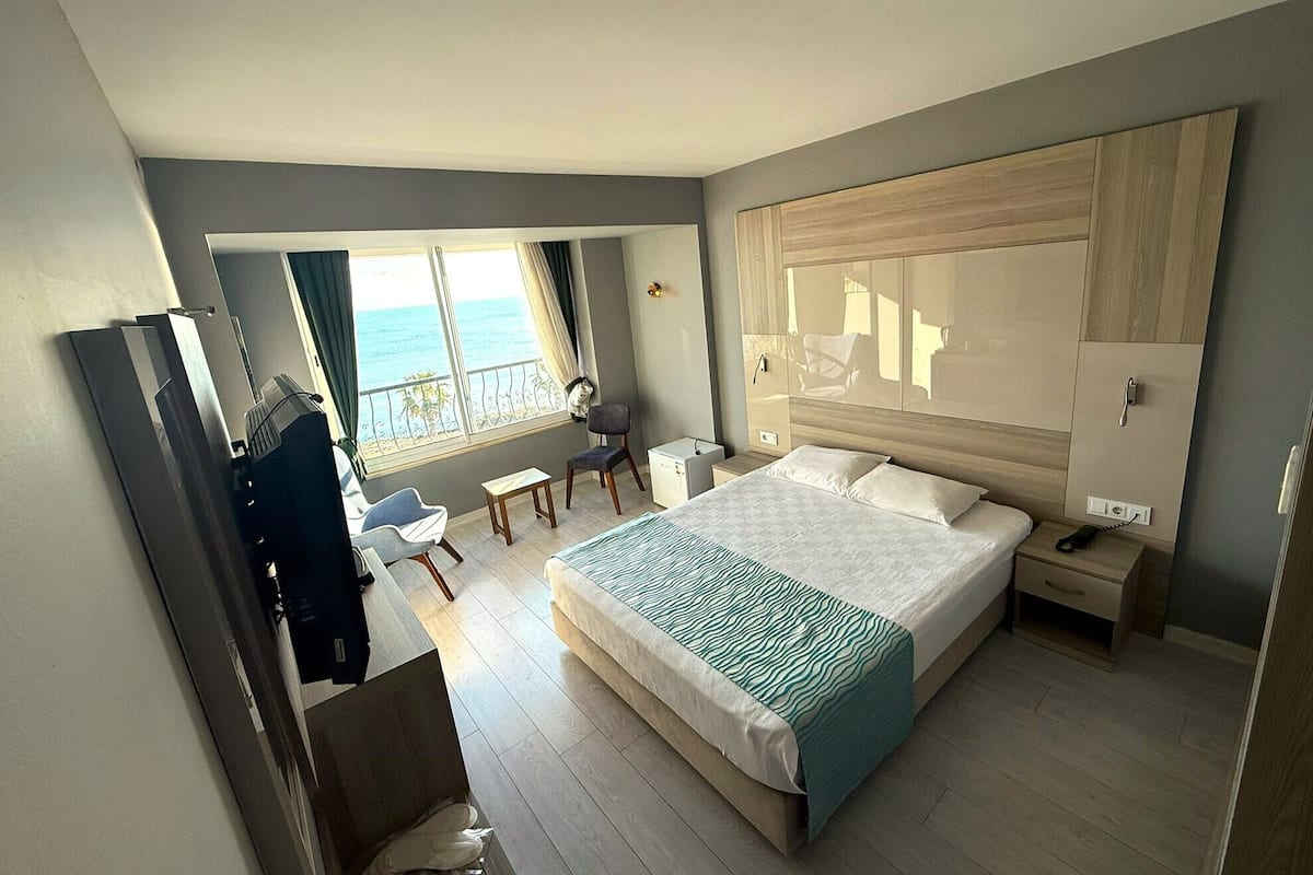 A modern hotel room with a large bed, a desk, a chair, and a view of the sea.
