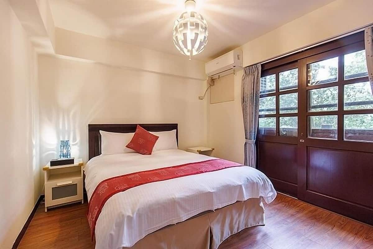 Quarto bem arrumado com cama, mesa de cabeceira e janela com cortinas.