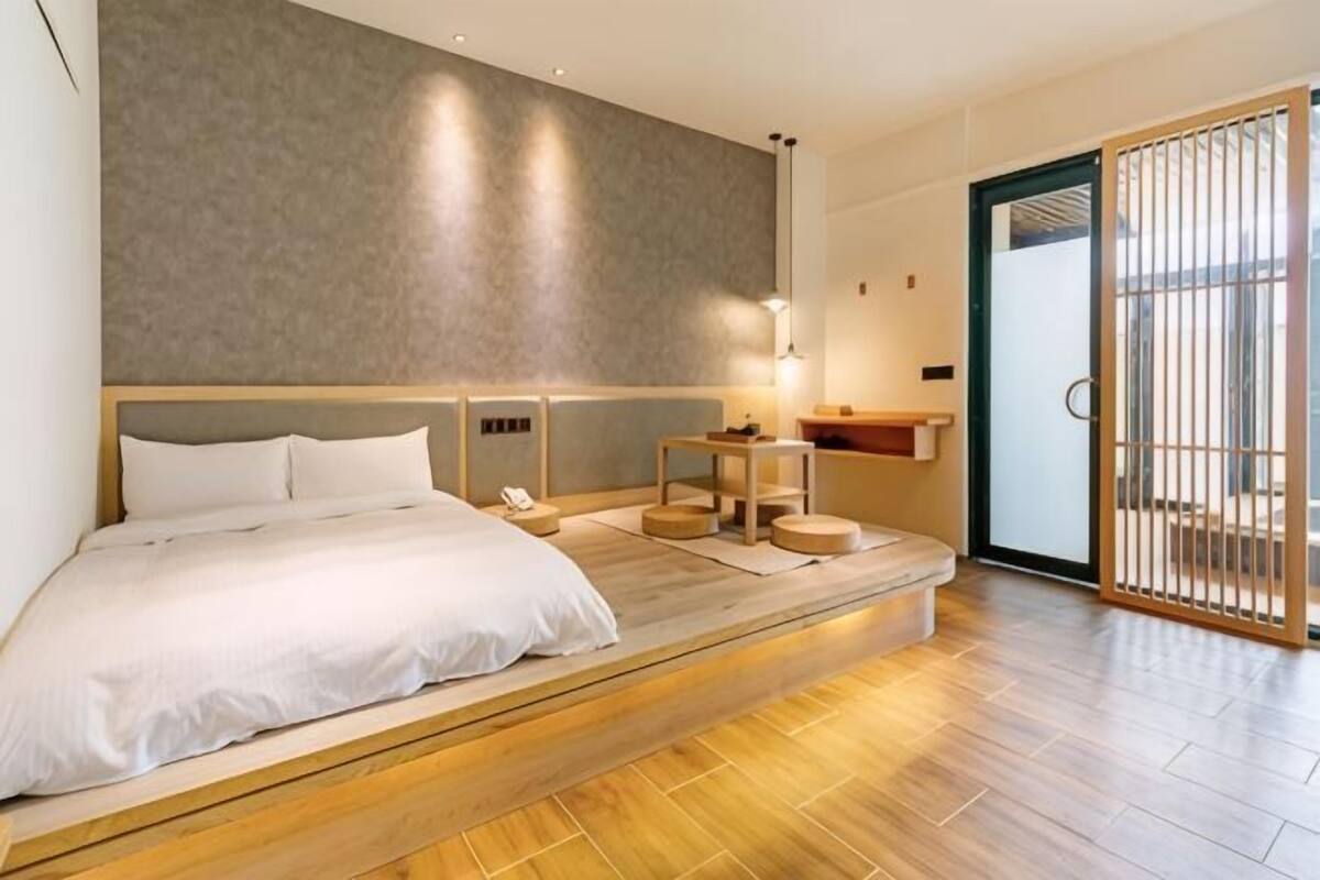 Quarto de hotel moderno com uma cama grande, uma mesa de cabeceira, uma pequena mesa e uma porta de vidro deslizante para o exterior.