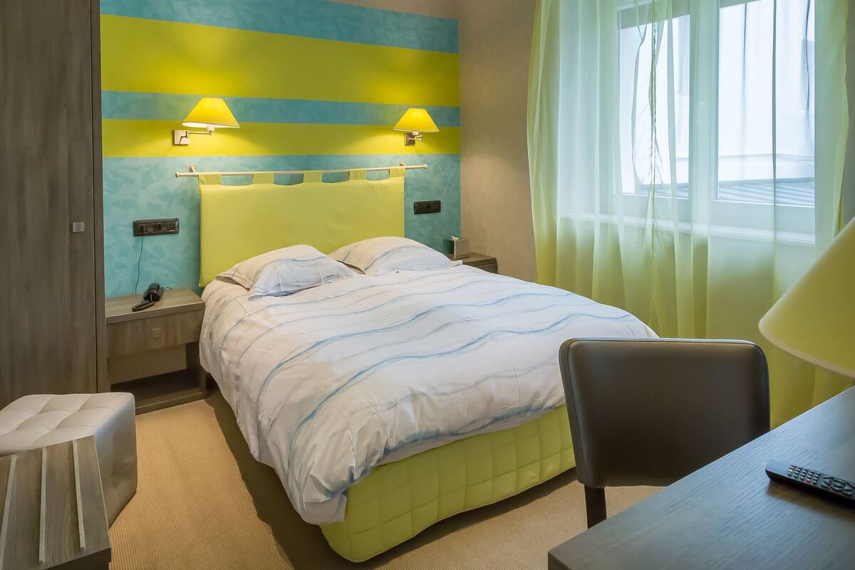 Ein Hotelzimmer mit Bett, Nachttischen, einem Sessel, einem Schreibtisch und einem fenster mit Vorhängen.