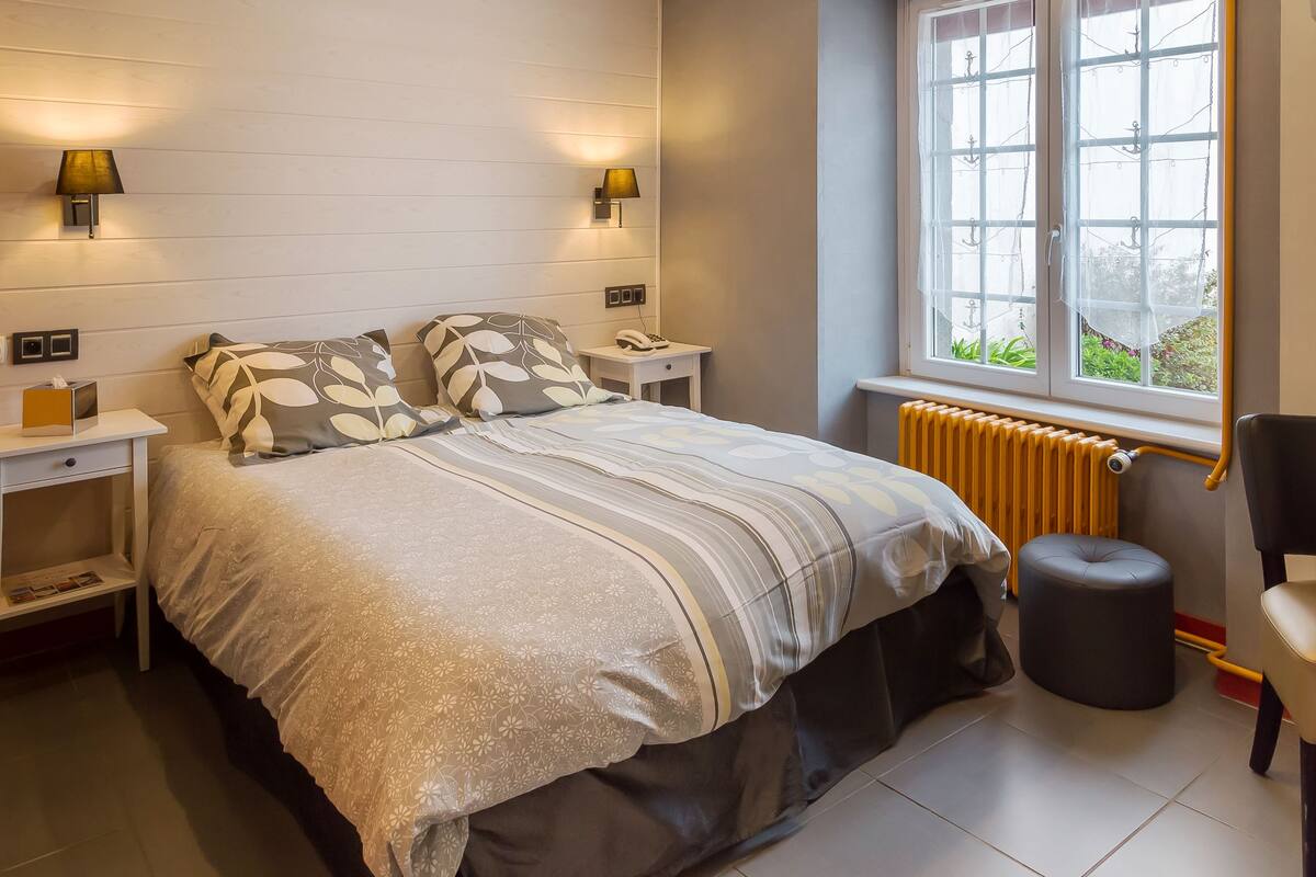 Ein Schlafzimmer mit Bett, Nachttischlampen, einem Nachttisch, einem Sessel und einem Fenster mit Blick ins Grüne.