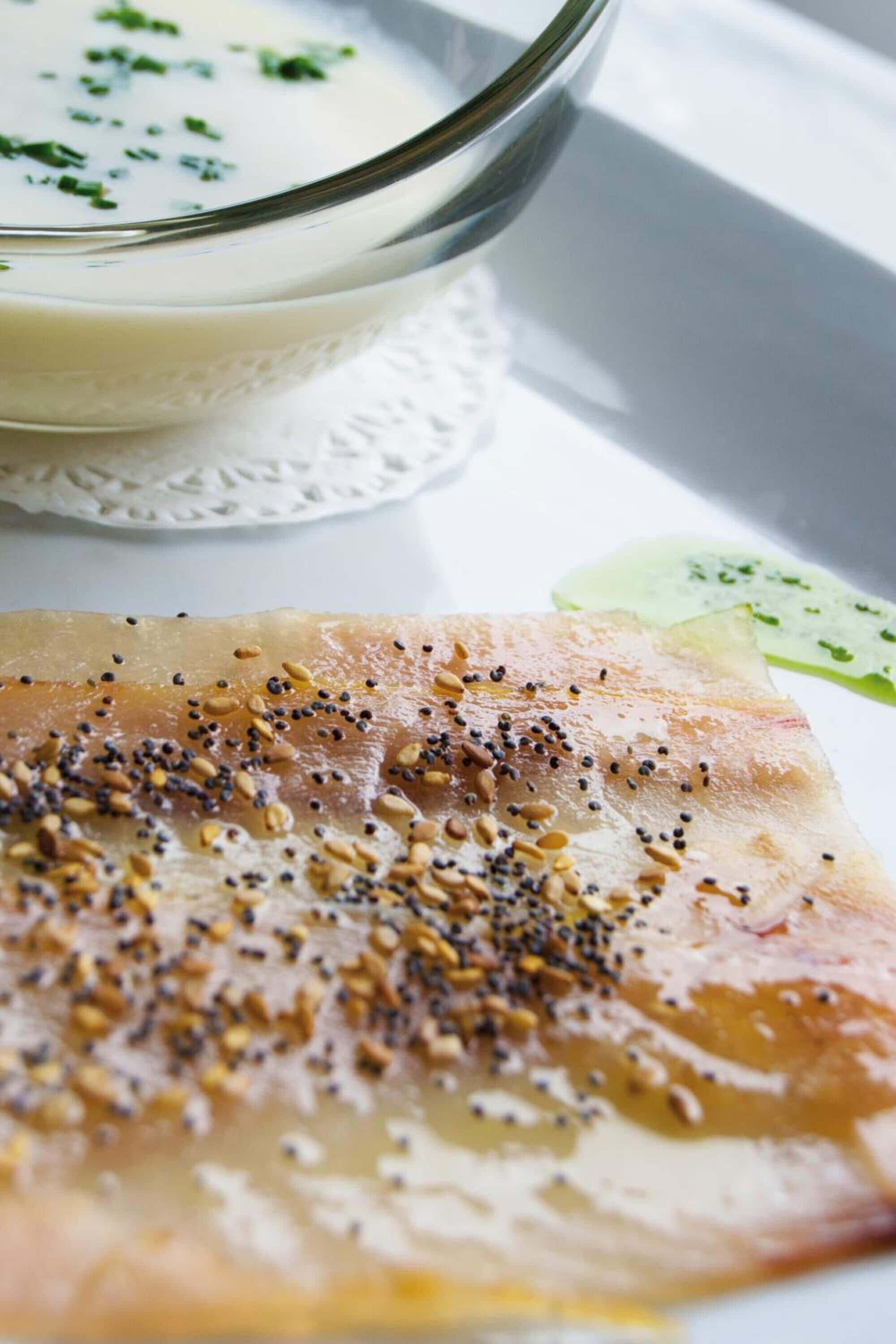 Un tazón de vidrio transparente con sopa y un adorno, una porción de pan plano con semillas y una rodaja de pepino.