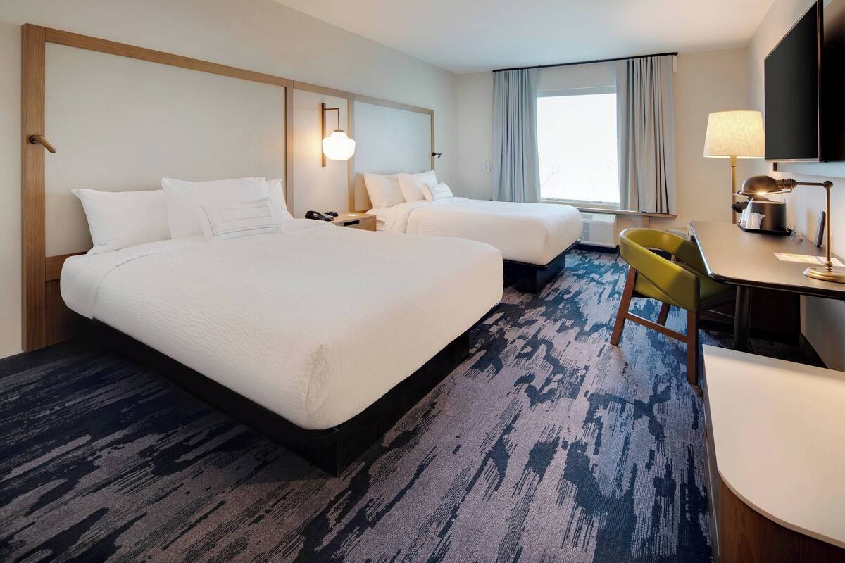 A hotel room with two beds, a desk, a chair, a TV, and a large window with curtains.