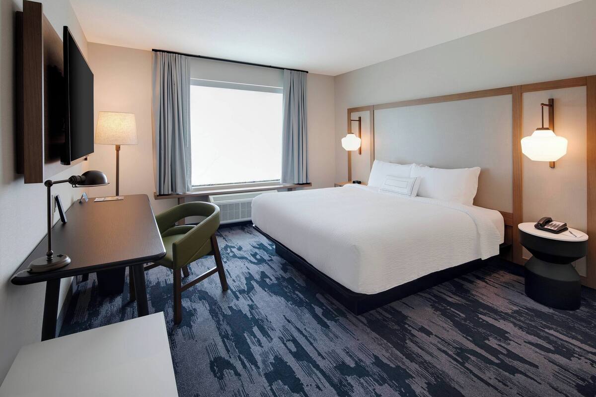 A hotel room with a bed, desk, chair, TV, lamp, and bedside table with a phone.