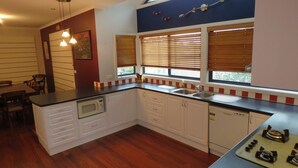 A compact kitchen with wooden cabinets, a microwave, a sink, and a stove. There are two windows with blinds, a dining area with a table and chairs, and a ceiling light fixture. - Central Holiday House at 29 Church in Cowes (Cowes)