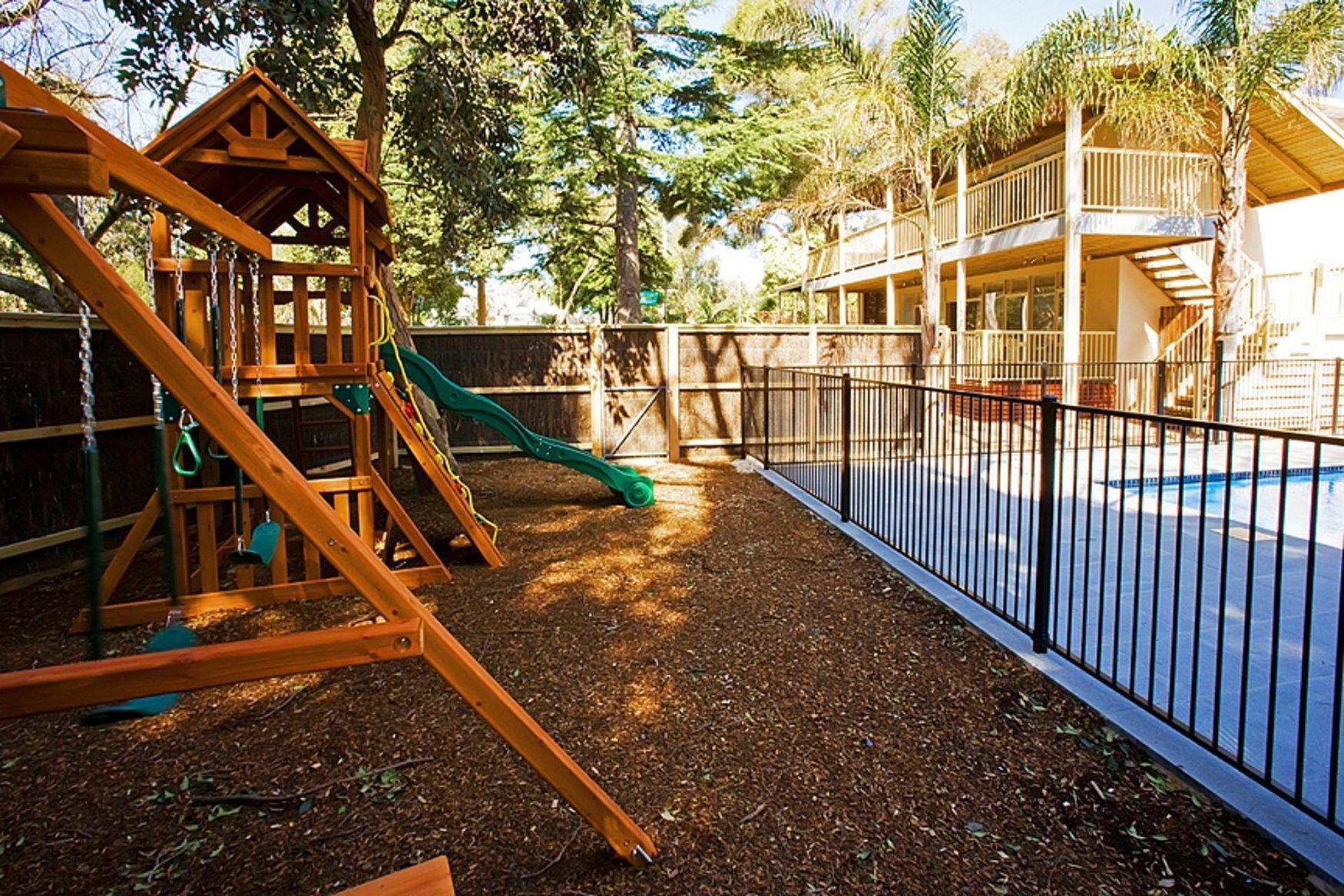 A playground with a wooden slide and swings, surrounded by a fenced area with a pool.