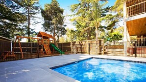 A swimming pool with a wooden play structure and a slide, surrounded by a fenced area with trees. - Beach House Units - 2 (Cowes)