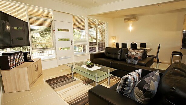 A modern living room with a glass coffee table, black leather sofa, and a television on a wooden cabinet. - Beach House Units - 3 (Cowes)
