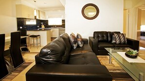 A modern living room with a black leather sofa, a glass coffee table, and a kitchen in the background. - Beach House Units - 3 (Cowes)