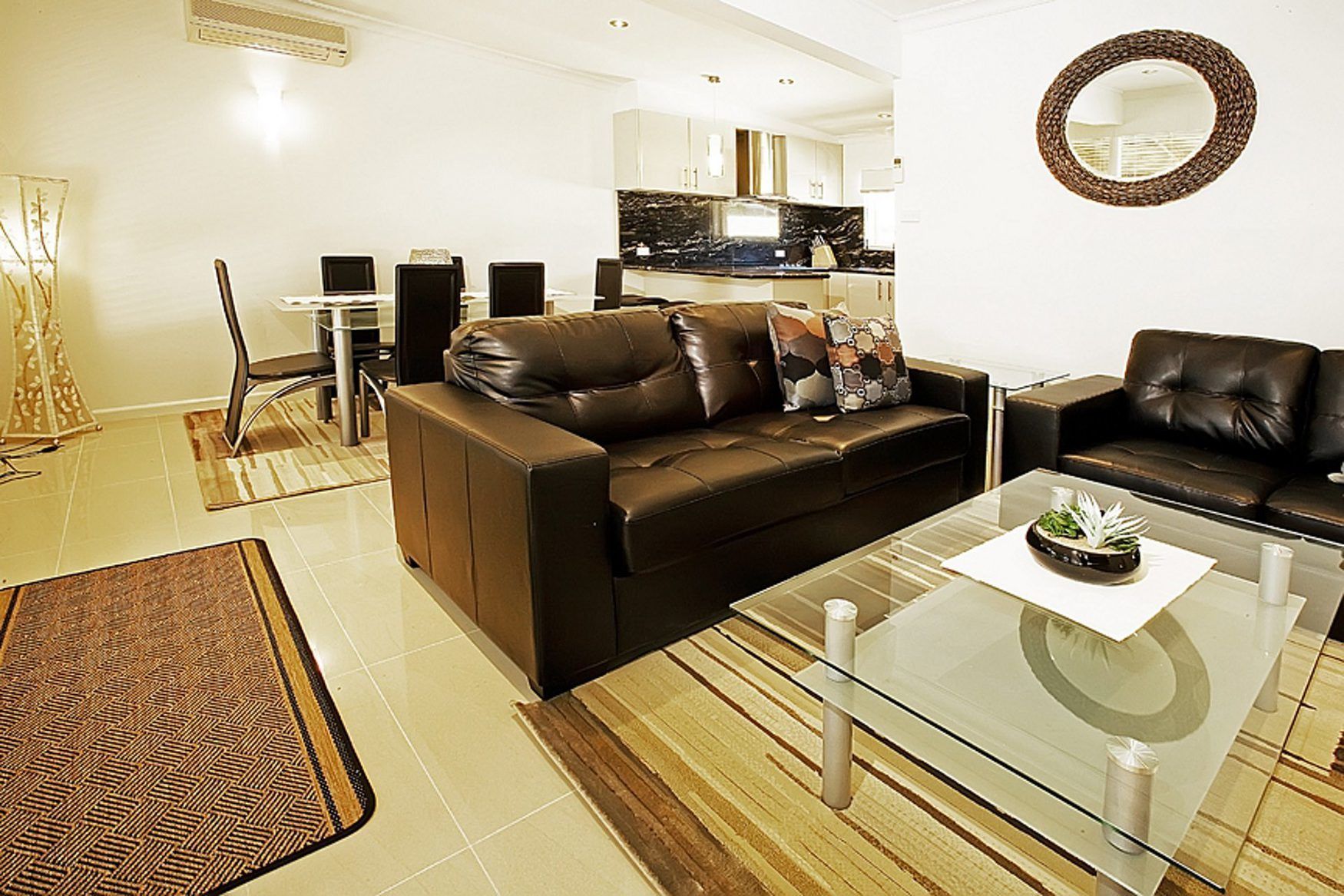A modern living room with a brown leather sofa, a glass coffee table, and a kitchen area in the background.