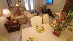 Una sala de estar con un sofá, sillas, una mesa de comedor dispuesta con platos y una planta, y un televisor en la pared.