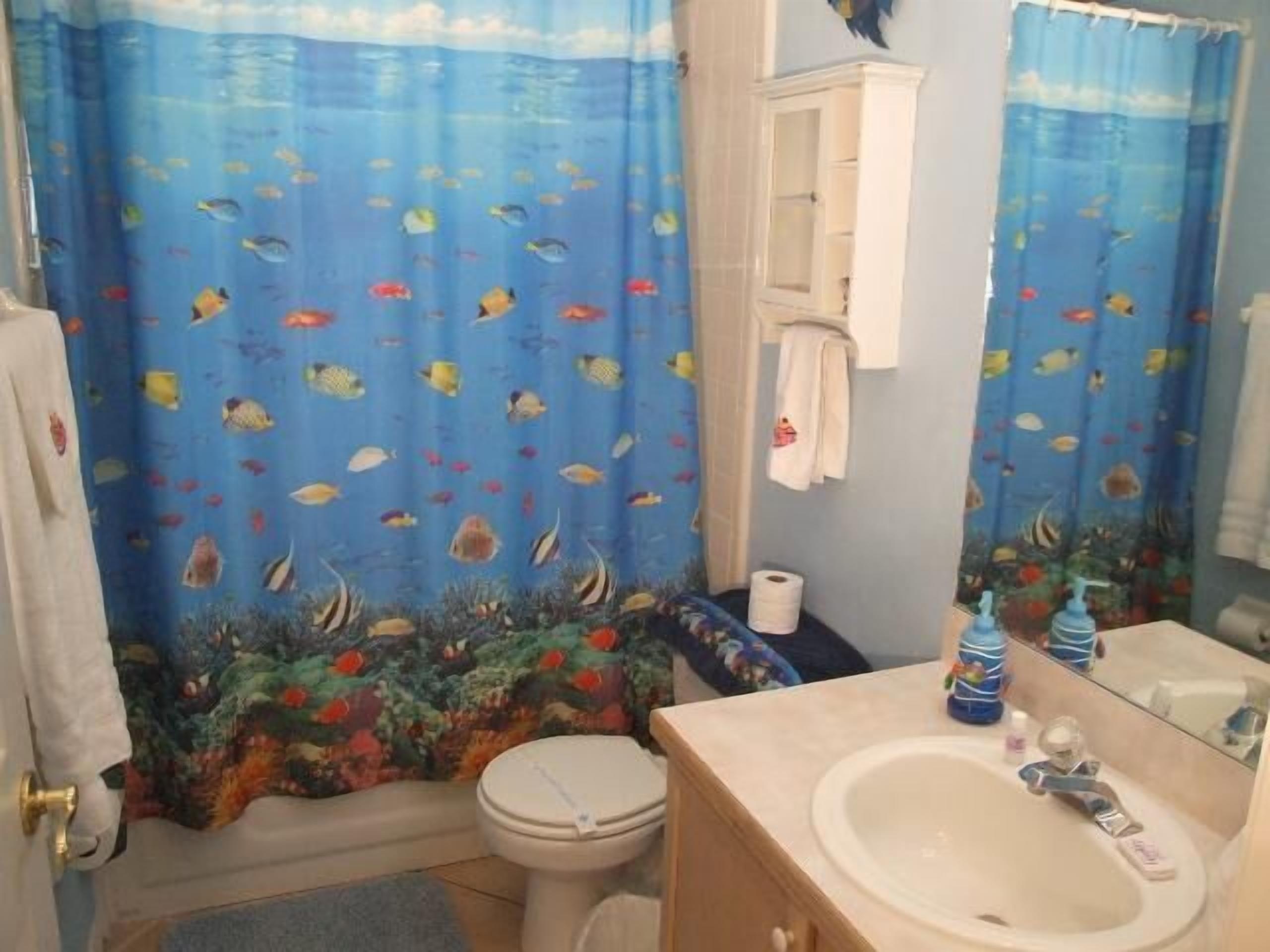 Et badeværelse med blå gardin med fiskemotiver, toilet, vask med spejl og skab.
