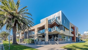 A modern multi-story building with a central atrium, outdoor seating area, and a palm tree in the foreground. - C-Scape by the Sea No.9 (Cowes)