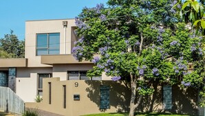 Ein modernes, zweistöckiges Haus mit beige-farbener Fassade, der Hausnummer 9 an der Vorderseite und einem Baum mit violetten Blüten im Vorgarten.