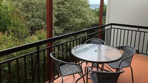 Varanda com mesa de vidro e cadeiras de vime, com vista para uma paisagem verdejante e um corpo d'água.