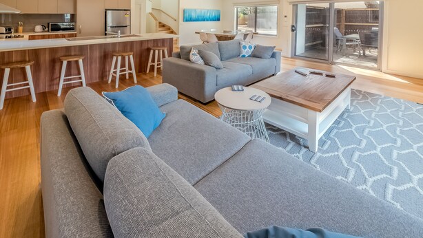 Una sala de estar moderna con un sofá gris, una mesa de centro y una zona de cocina con un refrigerador y taburetes de bar.