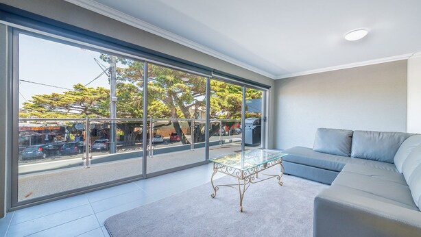 Ein modernes Wohnzimmer mit einem Glastisch, einem grauen Sofa und großen Fenstern mit Blick auf die Straße und Bäume.