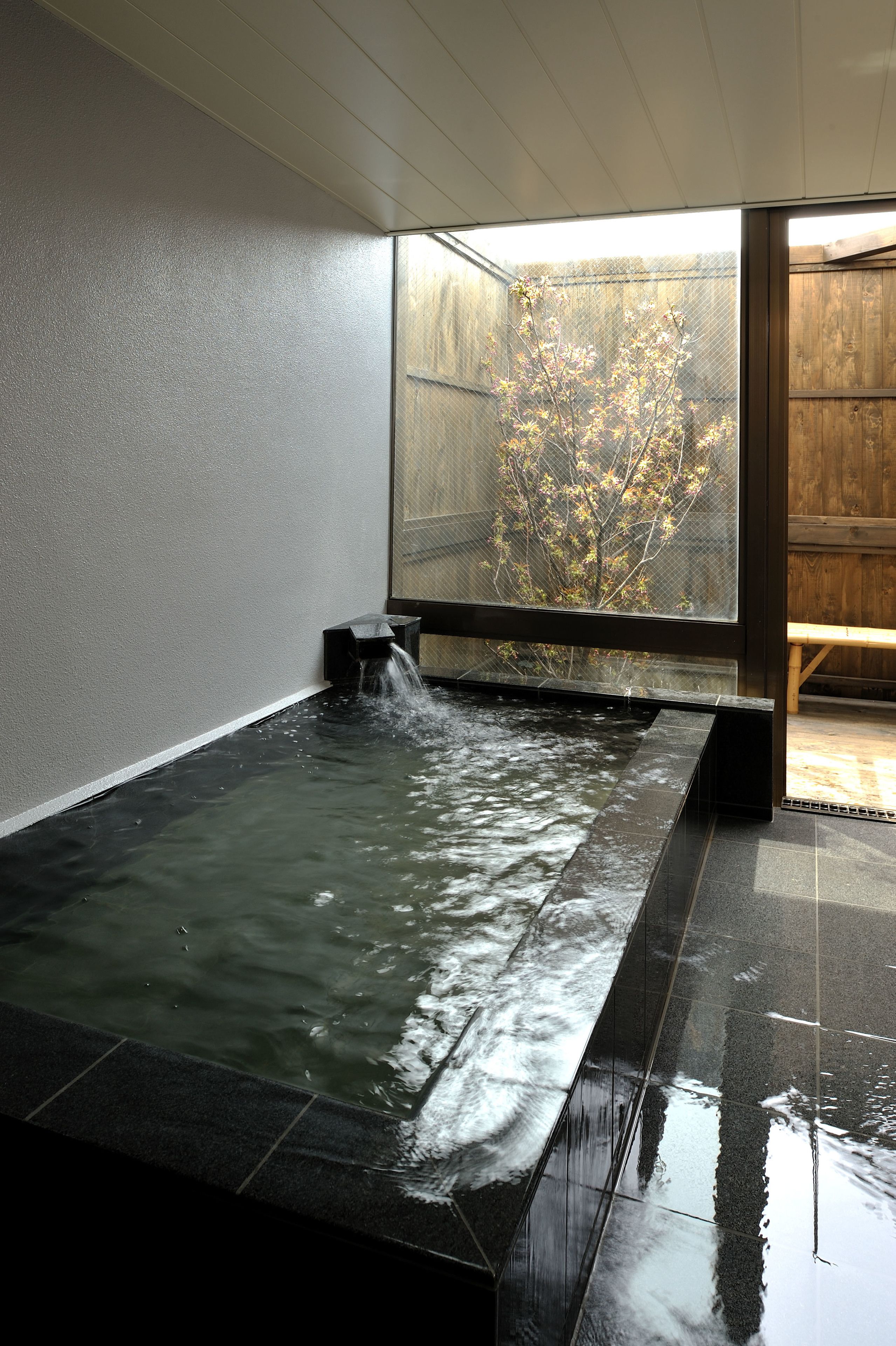A hot spring bath area with a wooden bench and a view of a tree outside.