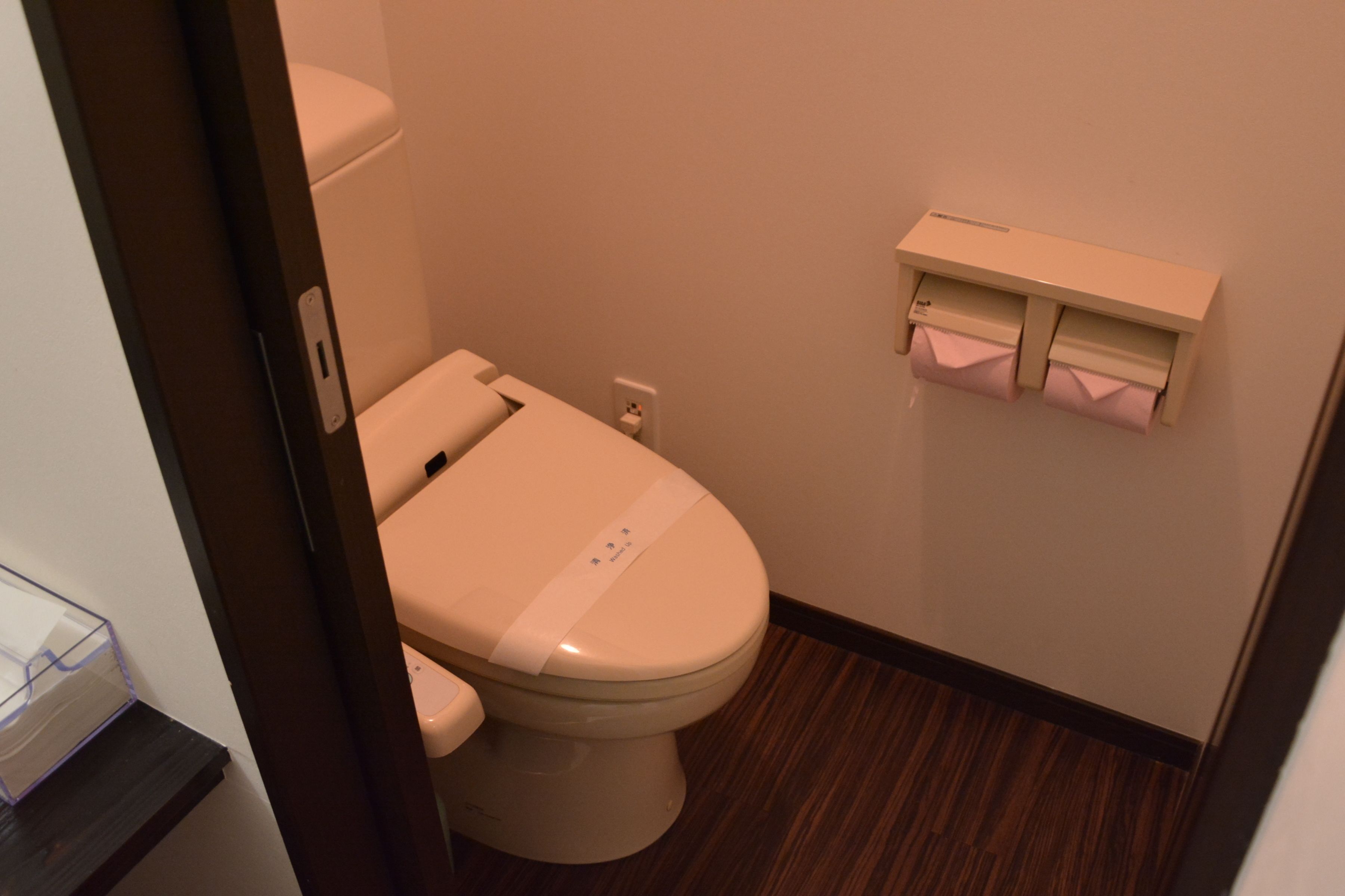 A small, compact bathroom with a toilet, a wall-mounted tissue dispenser, and a wooden floor.
