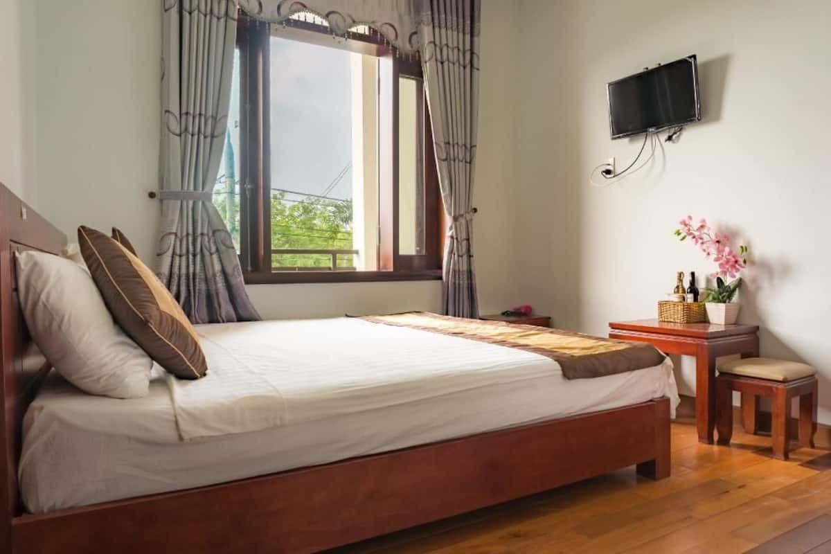 A neatly made bed with pillows, a wooden nightstand with a lamp, a small table with a vase of flowers, and a TV mounted on the wall.