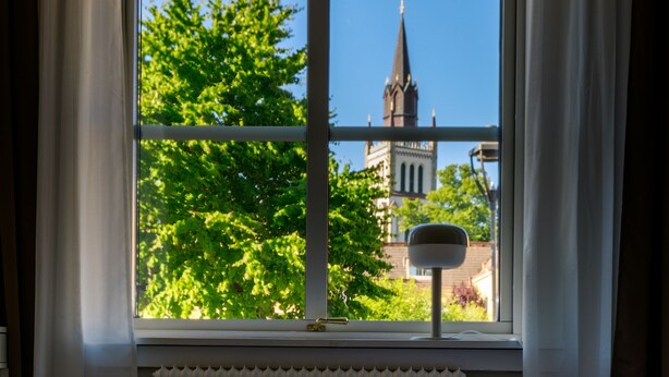 A room with a bed, a radiator, and a window with a view of a church and trees. - Vandrarhem Lidköping - Hostel (Lidköping)