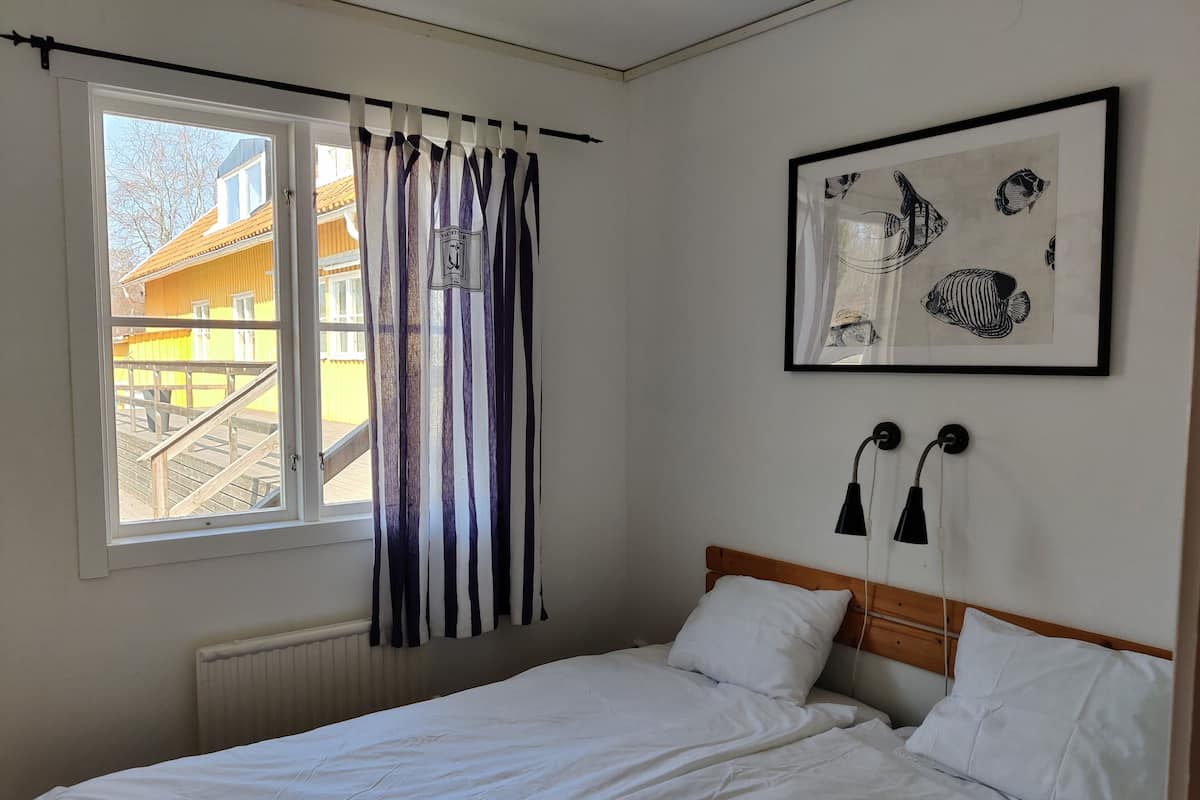 Une chambre à coucher comprenant un lit, une fenêtre avec des rideaux, un radiateur, un tableau encadré et deux lampes fixées au mur.