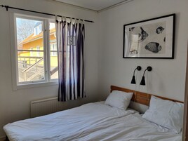 Un dormitorio con cama, ventana con cortinas, radiador, una obra de arte enmarcada y dos lámparas fijadas a la pared.