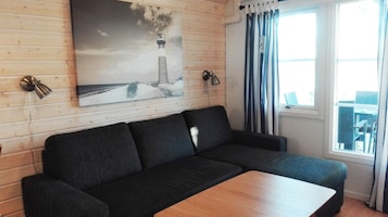 Un sofá oscuro con una mesa de madera al frente, un cuadro de un faro en la pared y una ventana con cortinas.
