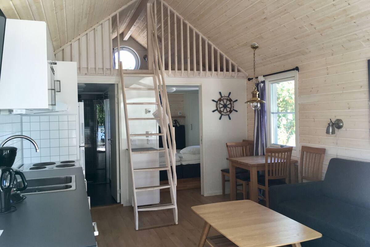 Un intérieur de chalet chaleureux, avec un revêtement en bois, une chambre à coucher située en mezzanine avec un puits de lumière, un coin repas et une cuisine équipée d’une cuisinière et d’un évier.