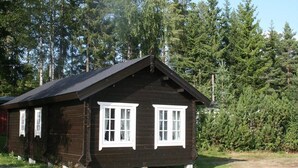 Eine kleine, dunkle Holzhütte mit weiß gefassten Fenstern und einem Satteldach, umgeben von dichtem Wald.