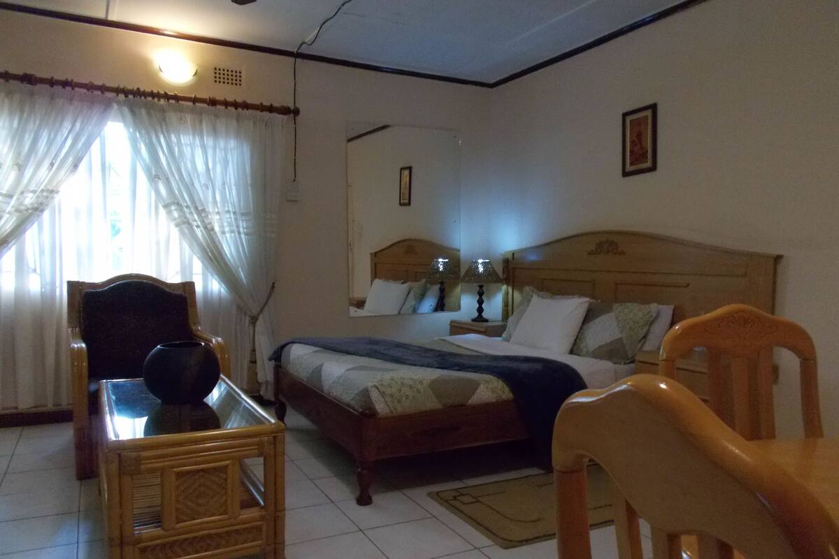 A bedroom with a bed, a wooden nightstand, a chair, a table, and a window with curtains.